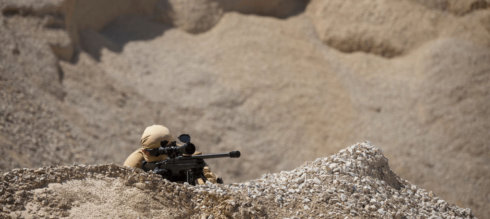 A person in beige tactical gear aims a sniper rifle in a desert-like environment, blending into the arid surroundings for camouflage.